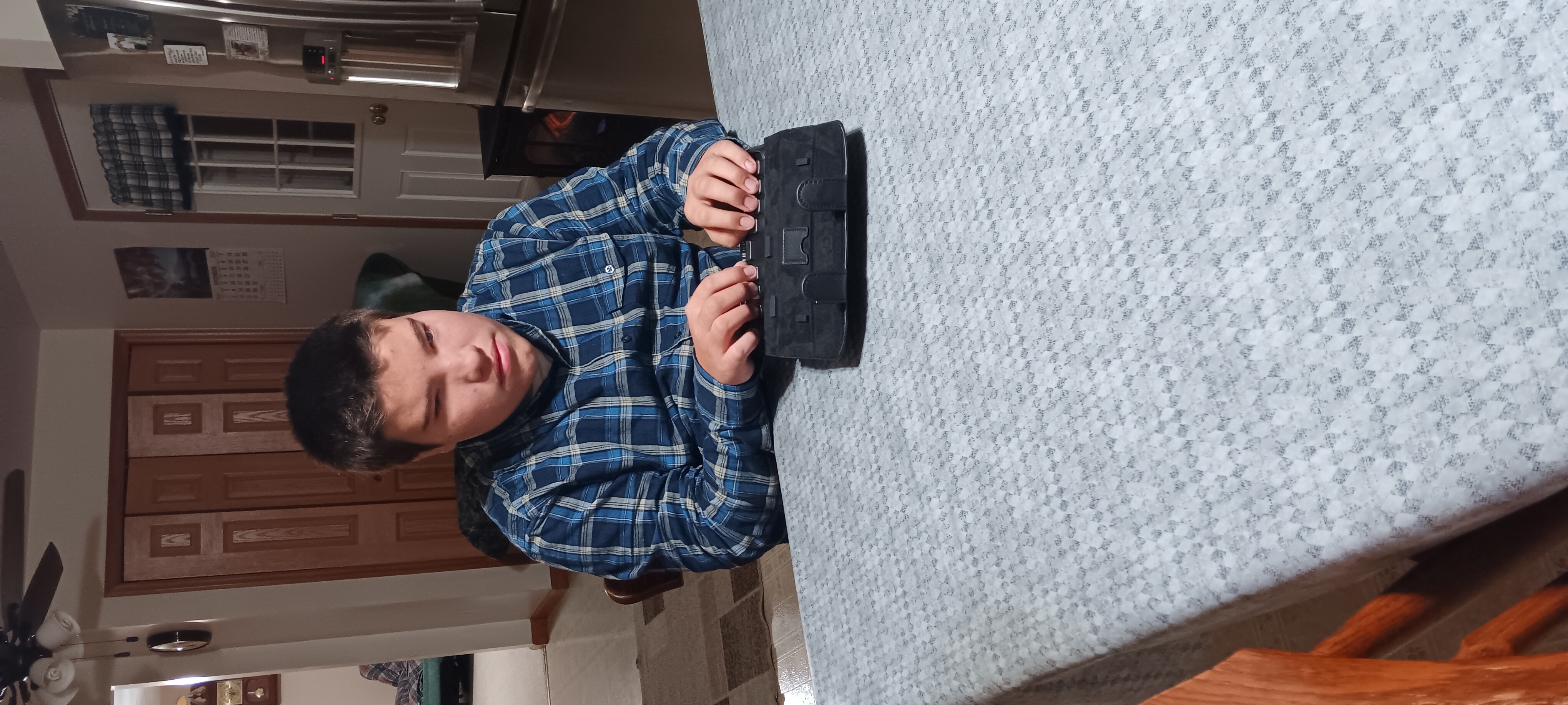 Wesley Martin, a young man wearing a blue and black plaid shirt, sits at a table using a Zoomax NLS eReader. The table is covered with a light-colored patterned tablecloth. The background shows a dining area with a ceiling fan, a refrigerator, a door with a window, and a calendar on the wall.